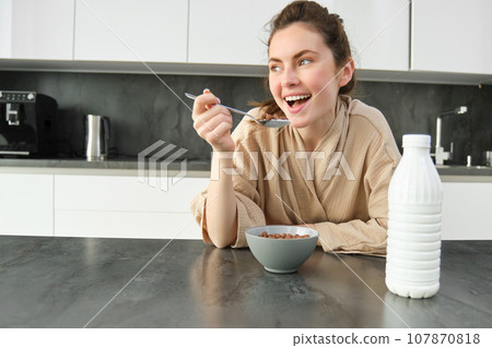 Portrait of young beautiful woman in bathrobe, eating cereals for breakfast, leans on kitchen worktop, looking at her morning meal Portrait of young beautiful woman in bathrobe, eating cereals for breakfast, leans on kitchen worktop, looking at her morning meal 107870818