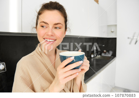 Portrait of beautiful young woman in bathrobe, drinking morning coffee and enjoying the taste, smiling pleased, standing in the kitchen 107870883