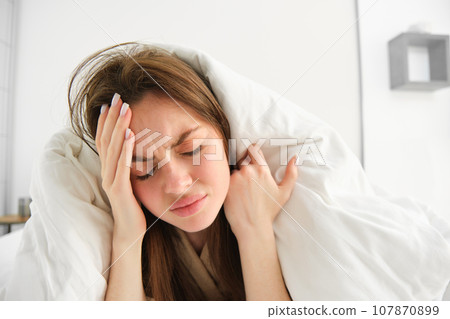 Close up of woman feeling unwell at home, lying in bed under white blankets and frowning, touching head, has headache, migraine, recovering from covid Close up of woman feeling unwell at home, lying in bed under white blankets and frowning, touching head, has headache, migraine, recovering from covid 107870899