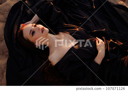 A woman in a black dress with a long train sits on a sand dune. A woman in a black dress with a long train sits on a sand dune. 107871126