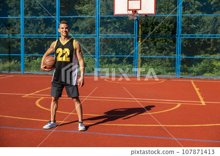 Athletic man standing on basketball court with ball underneath arm. 107871413