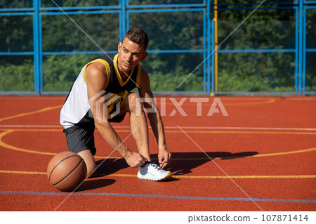 Athletic sporty man standing on basketball court ties his shoelaces before playing game. 107871414