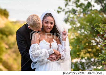 Newlyweds hug against the backdrop of a beautiful landscape, the guy kisses the neck of the girl who is looking into the frame Newlyweds hug against the backdrop of a beautiful landscape, the guy kisses the neck of the girl who is looking into the frame 107874503