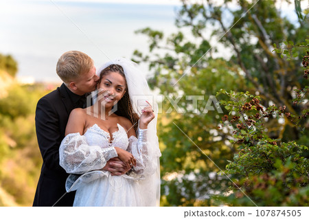 Newlyweds hug against the backdrop of a beautiful landscape, the guy kisses the girl who is looking into the frame 107874505