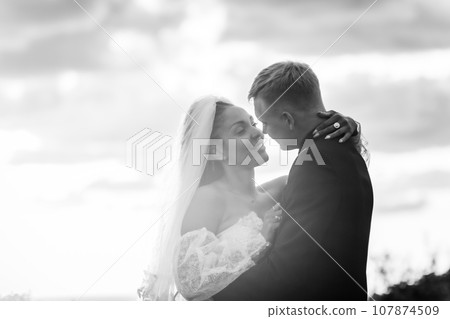 Happy newlyweds hugging against the sky in the rays of the setting sun, black and white version 107874509