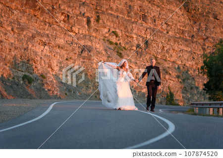 Happy newlyweds running along a mountain asphalt road 107874520
