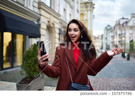 young woman in a red coat walks down the street and experiences joyful emotions looking at the smartphone in her hand. Good News, Winning, Discounts 107875786