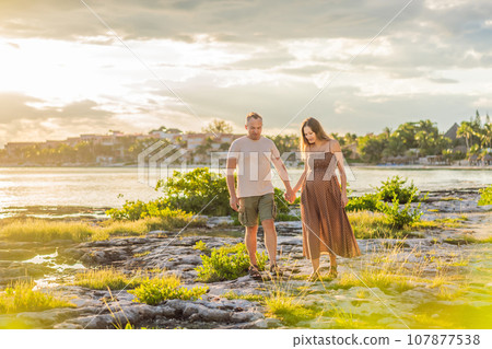 A happy, mature couple over 40, enjoying a leisurely walk on the waterfront On the Sunset, their joy evident as they embrace the journey of pregnancy later in life 107877538