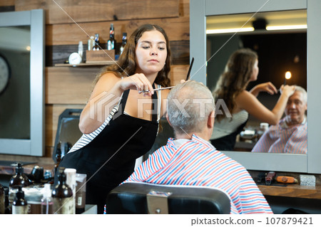 Girl master hairdresser completes creation of senior male clients image with help of scissors. 107879421