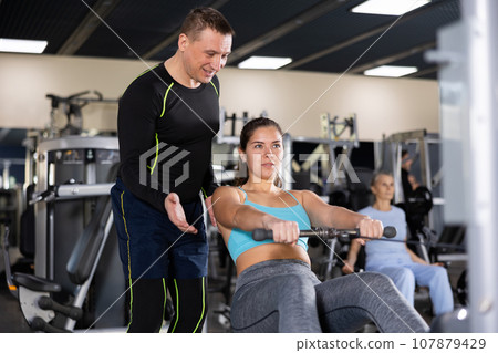 Male fitness instructor showing young woman how to use cable row machine 107879429