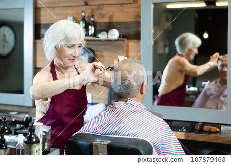 Experienced female hairdresser cutting hair of elderly client with scissors in barbershop 107879468