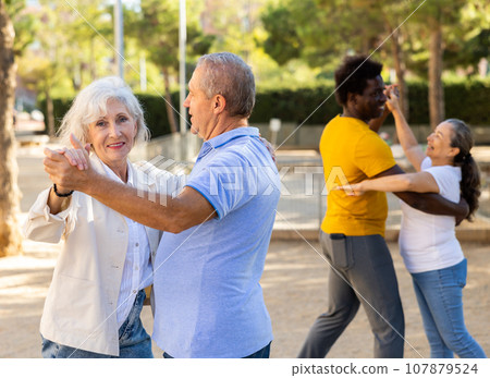 Exited older and middle-aged diverse people enjoying latin dances breaking into pairs on sandy area between trees 107879524
