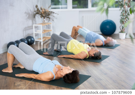 Old woman practicing bridge pose of yoga in training room during fitness classes Old woman practicing bridge pose of yoga in training room during fitness classes 107879609