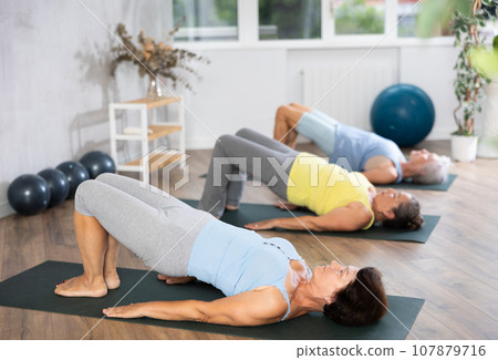 Old woman practicing bridge pose of yoga in training room during fitness classes 107879716