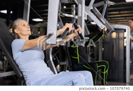 Aged woman performing chest press in exercise machine in gym 107879740