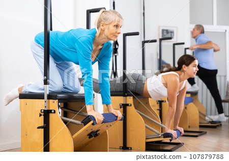 Focused mature woman performing stretching exercises while using pilates Wanda chair machine in fitness center Focused mature woman performing stretching exercises while using pilates Wanda chair machine in fitness center 107879788