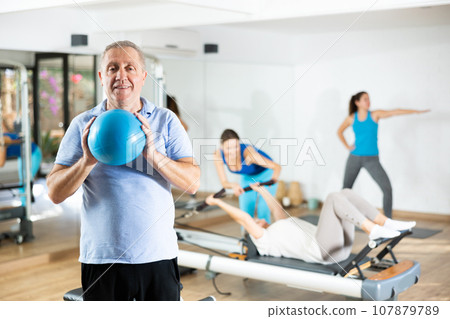 Smiling mature male squeezing ball in hands during Pilates training in gym Smiling mature male squeezing ball in hands during Pilates training in gym 107879789