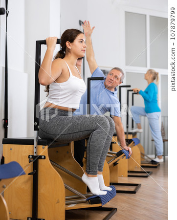 Calm positive young female in activewear concentrating on stretching exercise on Pilates chair equipment at gym. Active and healthy lifestyle rehabilitation concept 107879799