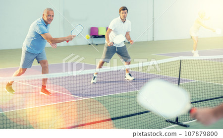 Two athletic men of different ages are playing a game of pickleball on a court inside sports facility 107879800
