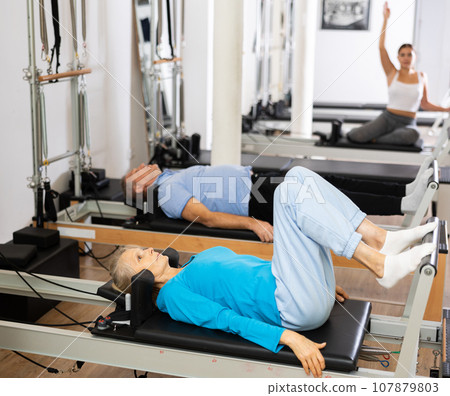 Focused elderly woman lying while doing Pilates tone exercise for back on reformer bed equipment in gym 107879803