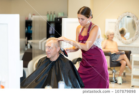 Young woman hairdresser performs haircut for elderly man using hair clipper Young woman hairdresser performs haircut for elderly man using hair clipper 107879898