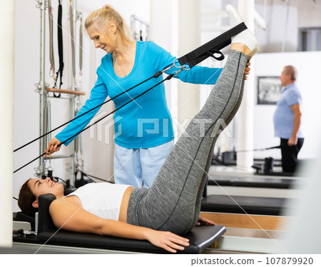 Professional mature personal female trainer assisting young woman exercising on Pilates machine in gym 107879920