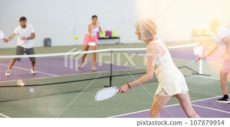 Rear view of aged woman playing pickleball on indoor court 107879954