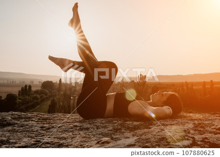 Fitness woman. Happy middle-aged fitness woman doing stretching and pilates on a rock near forest at sunset. Female fitness yoga routine. Healthy lifestyle with focus on well-being and relaxation. 107880625