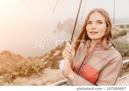 Woman rain umbrella. Happy woman portrait wearing a raincoat with transparent umbrella outdoors on rainy day in park near sea. Girl on the nature on rainy overcast day. 107880667
