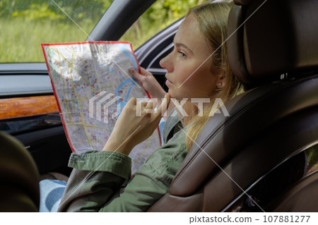 Blonde woman sitting inside the car checking the route on map to get to the destination. Afraid to get lost in forest. Young tourist explore local travel making candid real moments. True emotions 107881277