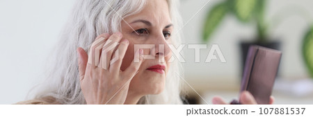Mature woman with long grey hair examines wrinkles on face 107881537
