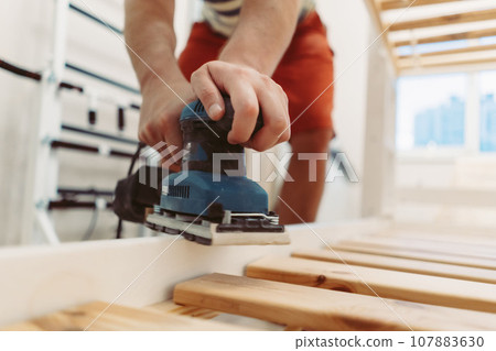 unrecognizable man rubs wooden child bed with power sander or grinder removing old paint unrecognizable man rubs wooden child bed with power sander or grinder removing old paint 107883630