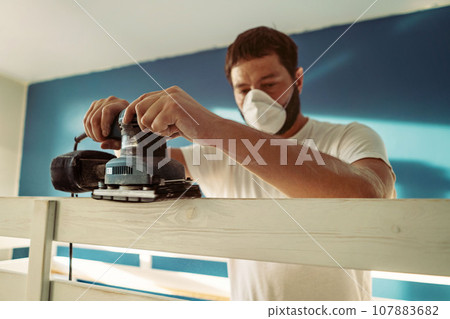 man rubs wooden child bed with power sander or grinder removing old paint man rubs wooden child bed with power sander or grinder removing old paint 107883682