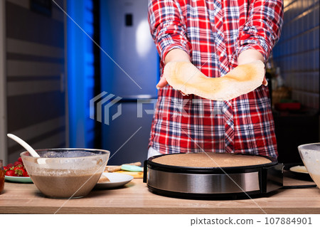 Woman cooking delicious crepe on electric pancake maker in kitchen, closeup 107884901