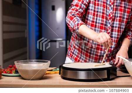 Woman cooking delicious crepe on electric pancake maker in kitchen, closeup 107884903