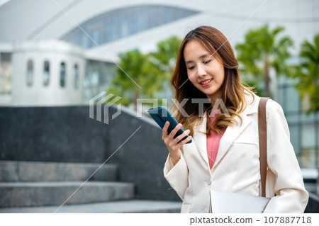 Portrait of confidence Asian businesswoman holding laptop and check message on mobile smart phone. She successfully work go to against city scene of modern office buildings in the city. cellphone 107887718