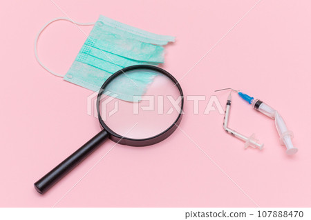 Broken Medical Syringe and Green Torn Medical Face Mask with Magnifying Glass 107888470