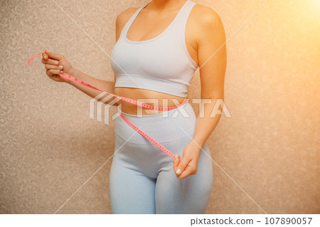 Cropped view of slim woman measuring waist with tape measure at home, close up. Unrecognizable european woman checking the result of diet for weight loss or liposuction indoors Cropped view of slim woman measuring waist with tape measure at home, close up. Unrecognizable european woman checking the result of diet for weight loss or liposuction indoors 107890057