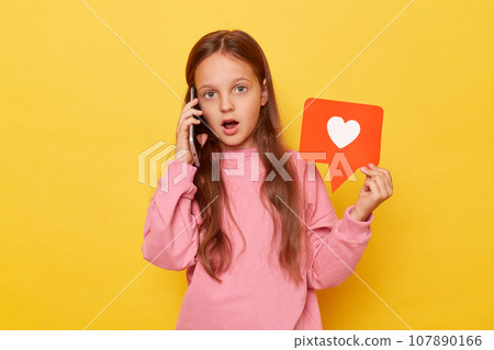 Shocked surprised amazed brown haired little girl, wearing casual clothing looking at camera with big eyes talking on ell phone showing like red icon sharing new by phone about her vlog and likes. Shocked surprised amazed brown haired little girl, wearing casual clothing looking at camera with big eyes talking on ell phone showing like red icon sharing new by phone about her vlog and likes. 107890166