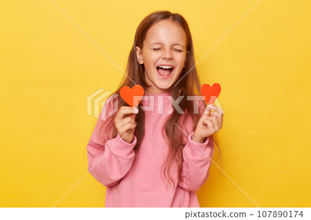 Overjoyed extremely happy brown haired little girl wearing pink sweatshirt isolated over yellow background standing with closed eyes and screaming or singing romantic song with red hearts in hands. 107890174