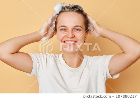 Attractive overjoyed woman washes hair applies shampoo taking shower standing with foam of head smiling happily isolated over beige background. Attractive overjoyed woman washes hair applies shampoo taking shower standing with foam of head smiling happily isolated over beige background. 107890217
