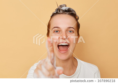 Beautiful cheerful joyful woman washes hair applies shampoo taking shower isolated over beige background showing foam on her finger click invisible button. Beautiful cheerful joyful woman washes hair applies shampoo taking shower isolated over beige background showing foam on her finger click invisible button. 107890218