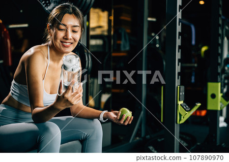 Asian girl holds green apple and water bottle in a gym, reminding viewers of the importance of balanced diet and hydration for achieving healthy lifestyle. Healthy fitness and eating lifestyle concept 107890970