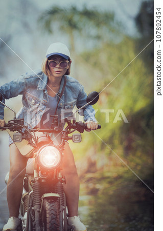beautiful woman wearing blue jeans jacket sitting on enduro motorcycle against colorful natural  background 107892454