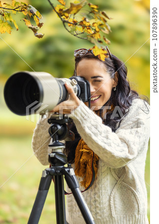 A satisfied photographer is standing behind her camera placed on a tripod, taking autumn photos A satisfied photographer is standing behind her camera placed on a tripod, taking autumn photos 107896390