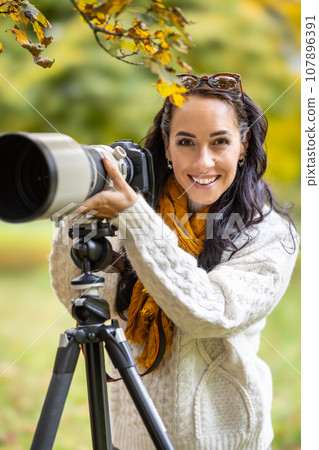 A professional female photographer stands next to her camera, taking pictures of autumn portraits or landscapes A professional female photographer stands next to her camera, taking pictures of autumn portraits or landscapes 107896391