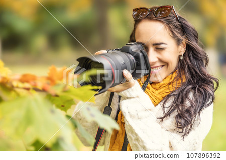 A smiling female photographer takes pictures of autumn leaves with her professional camera. 107896392