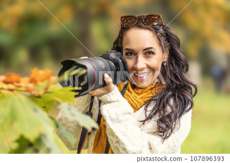 Beautiful woman holding a professional camera and looking for composition in autumn nature, she is smiling Beautiful woman holding a professional camera and looking for composition in autumn nature, she is smiling 107896393
