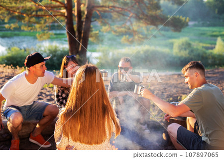 The company of girls and guys celebrate a holiday in nature. They clink glasses with drinks. Rest in the summer in the forest on the banks of the river against the backdrop of sunset. Tourism and 107897065