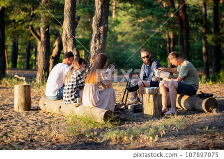 A company of friends of girls and guys have a rest in the summer in a picturesque place in the forest on the banks of the river against the backdrop of sunset. Picnic in nature, tourism. Cooking tea A company of friends of girls and guys have a rest in the summer in a picturesque place in the forest on the banks of the river against the backdrop of sunset. Picnic in nature, tourism. Cooking tea 107897075